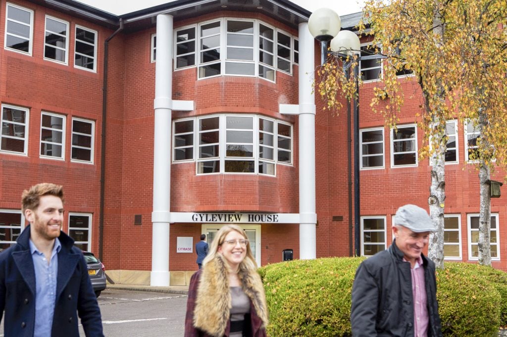 A red-brick building labeled Gyleview House, nestled in Edinburghs charm, features large windows and white accents. Three people stroll by, smiling beneath the overcast sky. To the right, a tree adorned with autumn leaves adds warmth to the scene.