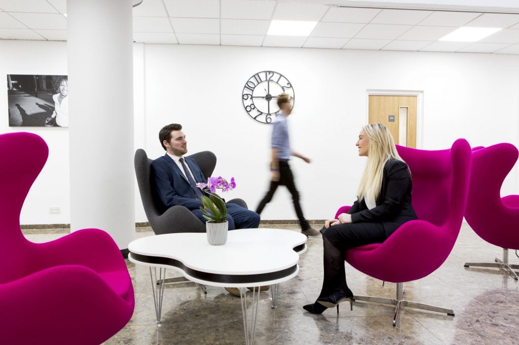 Two people in business attire sit on modern pink chairs around a white coffee table with a plant, reminiscent of an Edinburgh Gyleview office. A person walks by in the background. The room features a wall clock and a black-and-white photo on the wall.