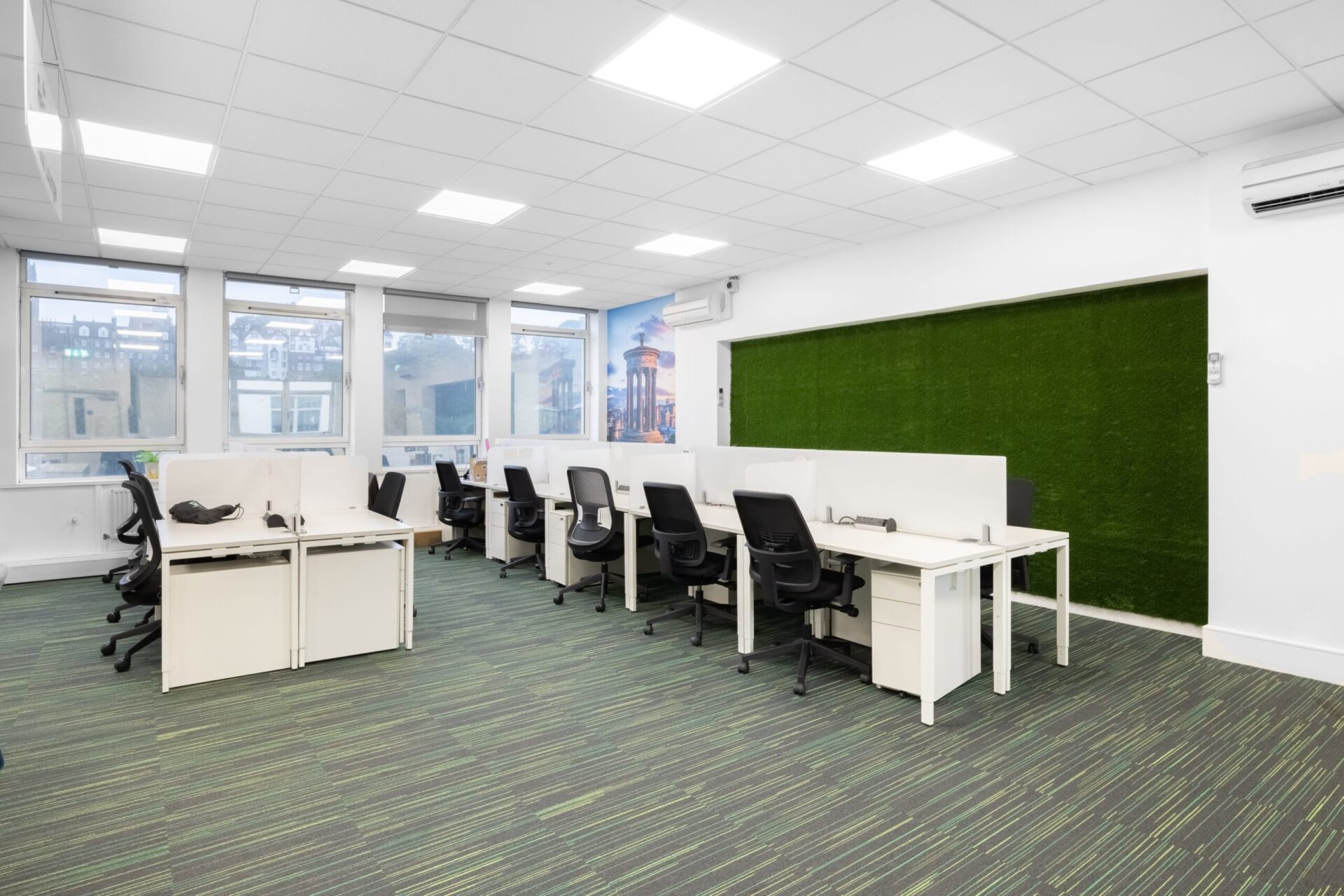 A modern office space on Princes Street features white desks and black chairs arranged in rows. Partitions divide the desks, and the green-patterned carpet complements the natural light pouring through large windows, with a striking green accent panel adding a touch of style.