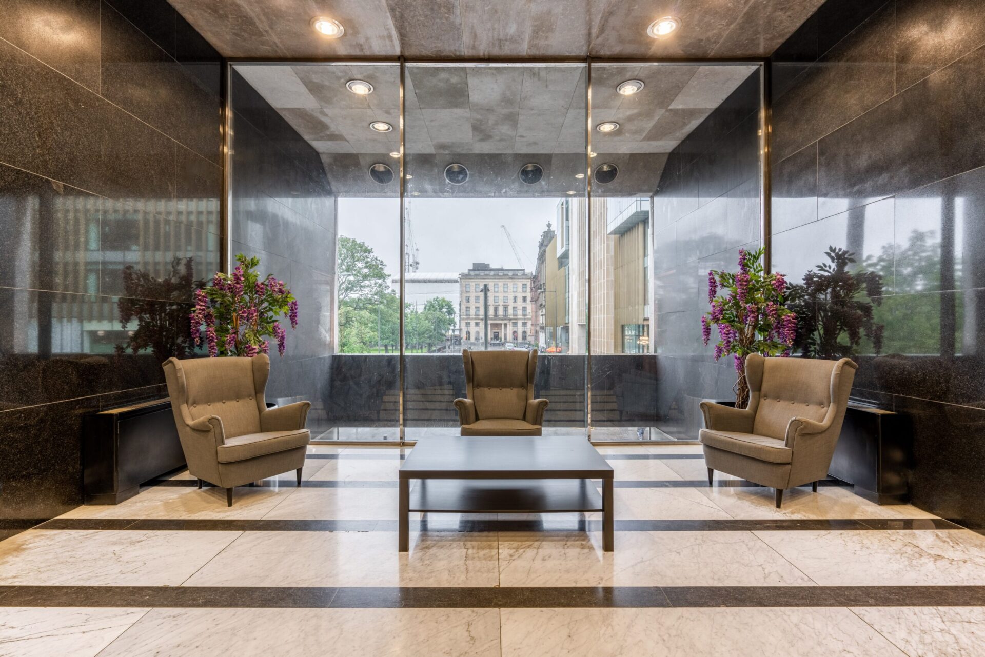 The spacious modern lobby at St Andrew Square features three beige armchairs and a rectangular coffee table on a striped marble floor. Large glass doors offer a view of trees and nearby buildings, complemented by elegant floral arrangements on either side.