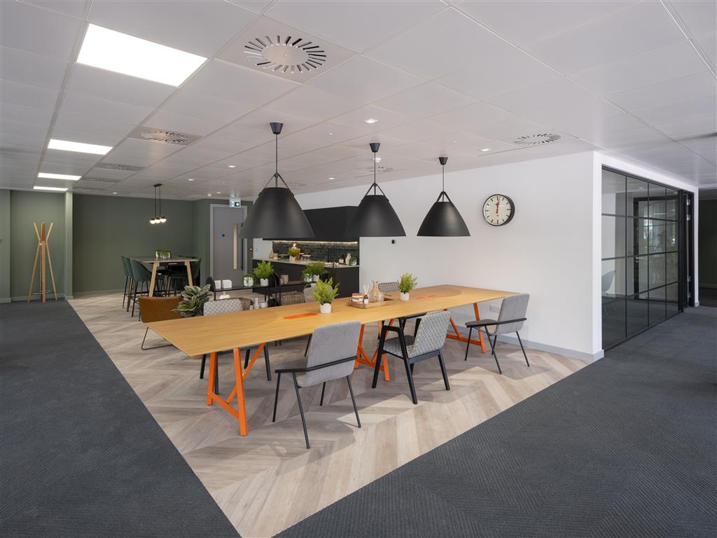 A verdant touch graces the modern office break area, featuring a long wooden table, six chairs, black pendant lights, and a wall clock. Lush plants enhance the space while a small kitchen with light green walls and wooden flooring is visible in the background.