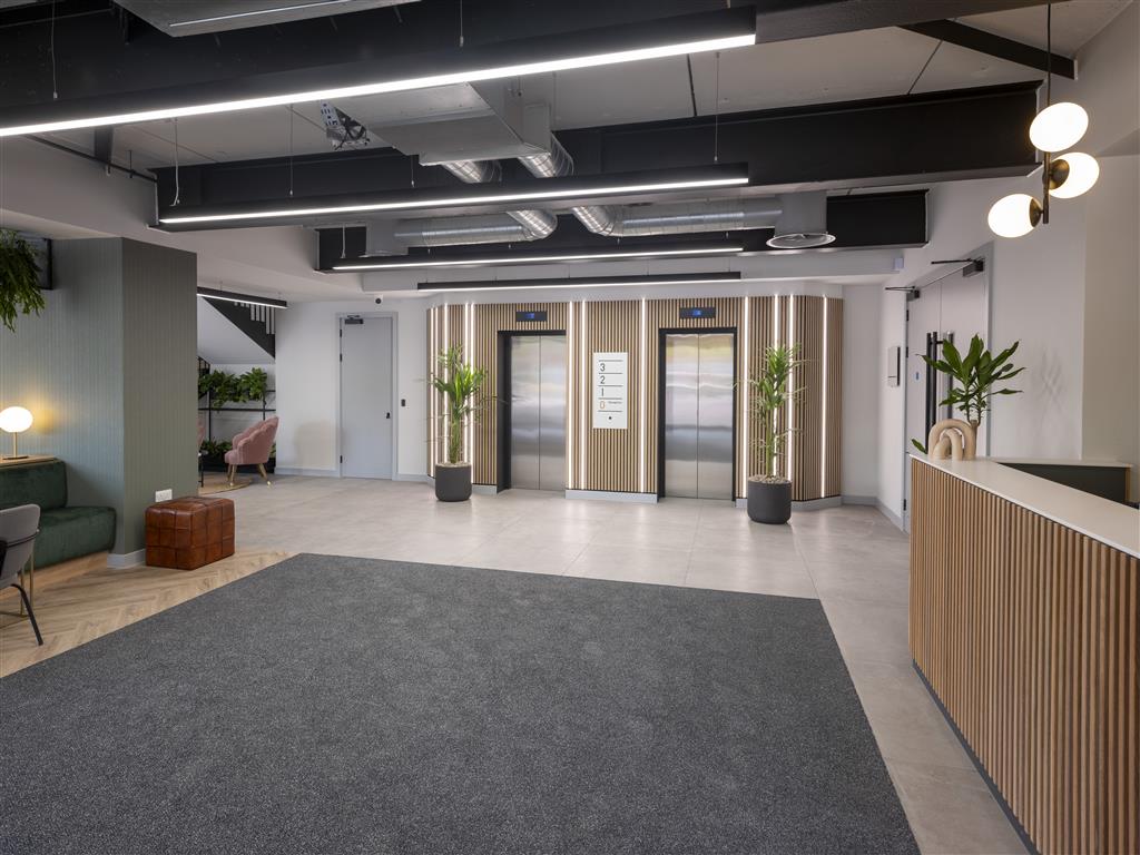 A modern office lobby with two elevators on a verdant gray carpet and a sleek wooden reception desk to the right. The ceiling showcases exposed ductwork and linear lights, while potted plants offer lush greenery near the elevators and in corners.
