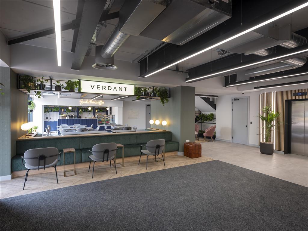 The modern office lobby showcases sleek furniture beneath signage reading Verdant above the reception desk. Potted plants enhance the space, complementing the industrial ceiling and elevator. In the background, a seating area rests on a blend of carpet and tiles.
