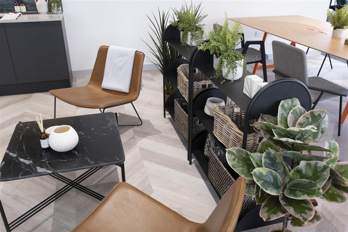 Modern living space with brown leather chairs, a black marble coffee table, and a dark shelving unit adorned with verdant plants and wicker baskets. Wooden dining table in the background, light wood flooring, and a white wall enhance the airy atmosphere.
