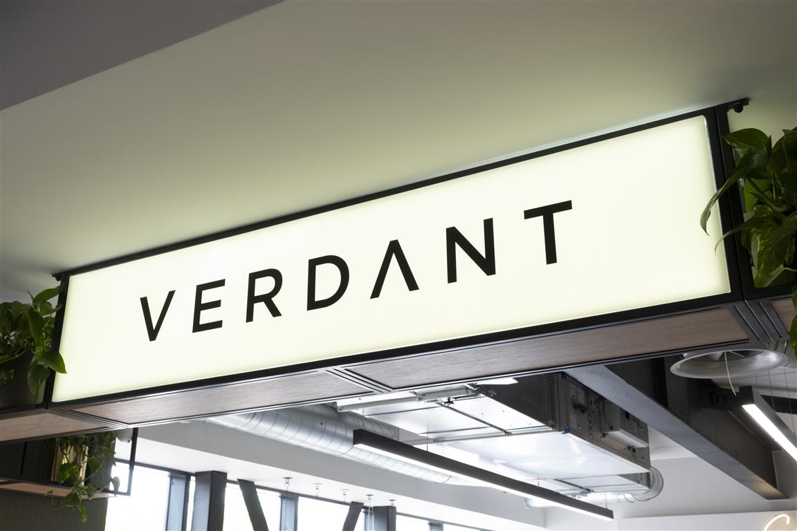 The illuminated sign, boldly displaying the word VERDANT in striking black letters, is embraced by lush green plants. This vibrant centerpiece complements a modern interior accentuated by exposed ductwork and sleek gray beams.