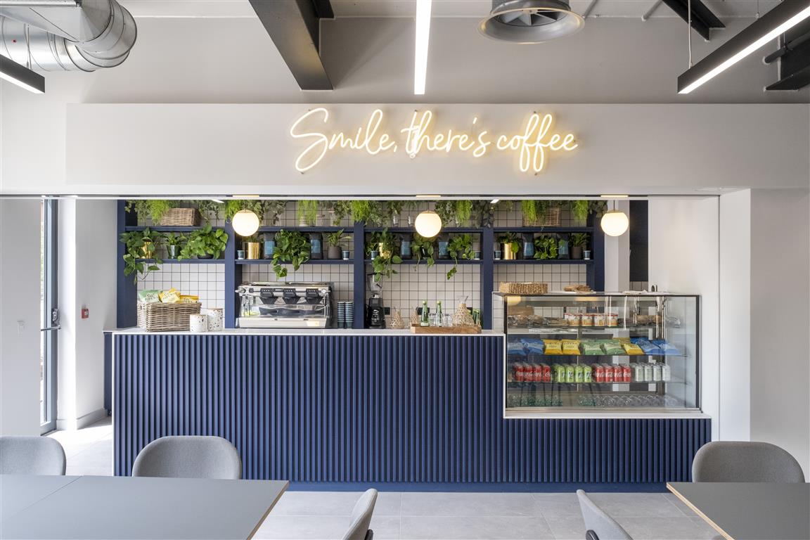A modern café interior with a blue counter and a neon sign reading Smile, theres coffee. The counter hosts a coffee machine, pastries, and snacks. Verdant potted plants adorn the shelves above. Gray chairs and tables are in the foreground, creating an inviting atmosphere.