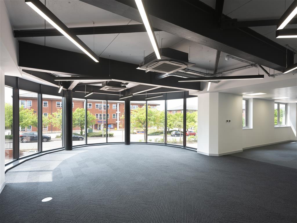 Spacious, modern office interior with large floor-to-ceiling windows, black carpet, and angular ceiling lights. Outside the windows, a view of parked cars and a brick building lined with verdant trees is visible.