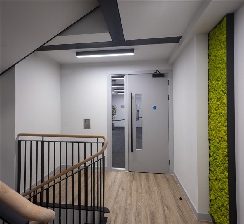 A modern hallway with wood flooring showcases a verdant moss panel on the right wall. A backlit staircase railing glows on the left, guiding you toward a gray door with two vertical windows straight ahead. Overhead, a fluorescent light illuminates the space.