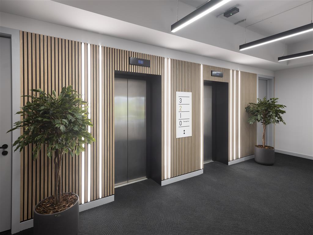 A modern elevator lobby features three elevators framed by vertical wooden slats. Verdant potted plants grace each end, while a floor directory is centered above the middle elevator. The space is well-lit with linear ceiling lights, creating an inviting atmosphere.