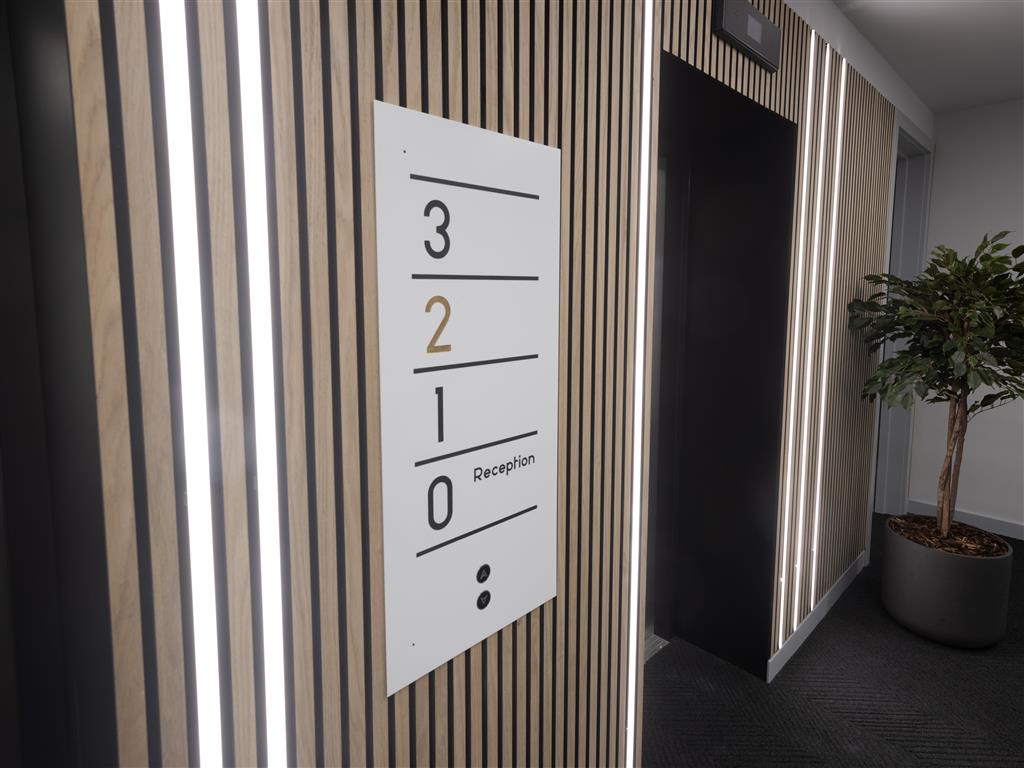 A modern hallway features vertical wooden slats on the wall. A white sign displays floor numbers in braille: 3, 2, 1, and 0 labeled as Reception. A verdant potted plant graces the right side, while lights warmly illuminate the area.