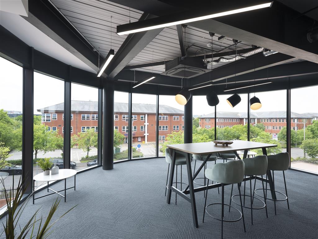Modern office space with large floor-to-ceiling windows offers a verdant view of neighboring buildings and lush trees. Inside, a tall table with five bar stools and contemporary light fixtures create a sleek, open atmosphere.
