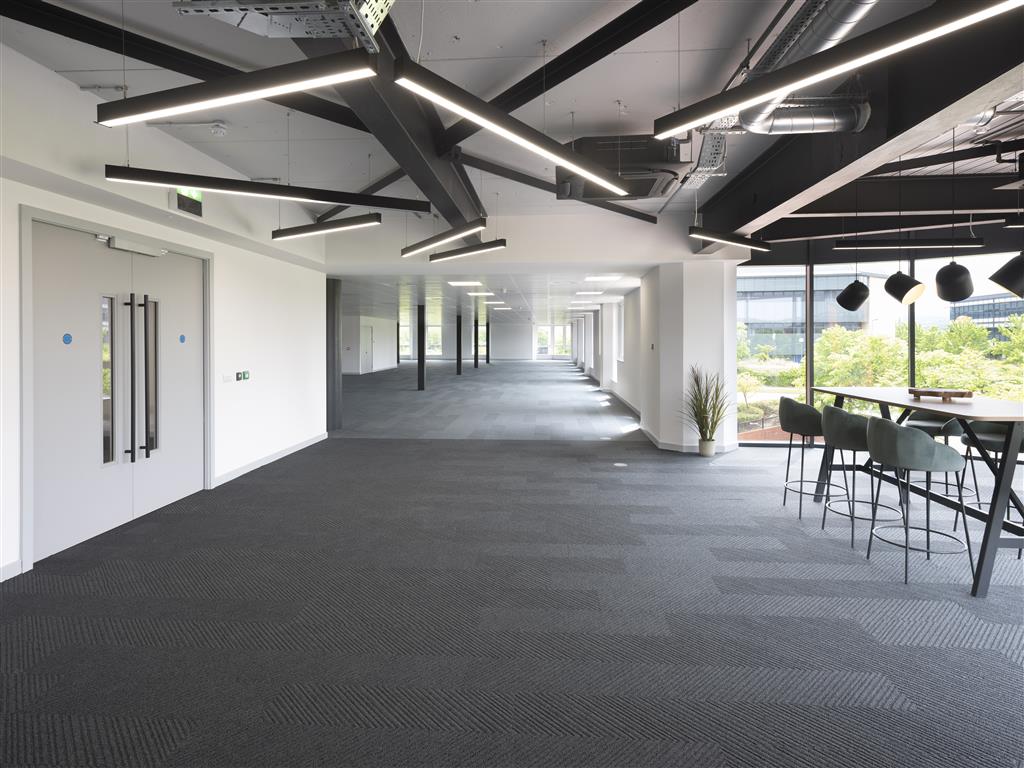 Spacious modern office space with a verdant touch, enhanced by dark carpet and angular ceiling lights. Large windows line the right wall, inviting natural light. A bar table with stools stands near the windows, while an exit door is visible on the left.