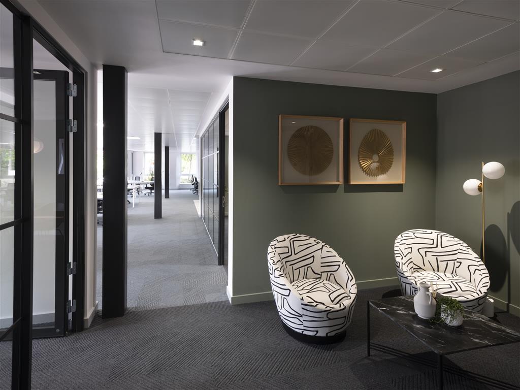 The modern office interior boasts a verdant feature wall adorned with two abstract artworks. Two patterned chairs flank a sleek black coffee table, complemented by a minimalist lamp. Glass partitions subtly separate the office space in the background.
