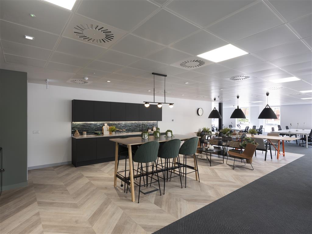 Modern office space with a kitchenette featuring dark cabinets and a high table surrounded by verdant green chairs. Pendant lights hang above, illuminating an open seating area with white desks. Large windows flood the space with natural light.