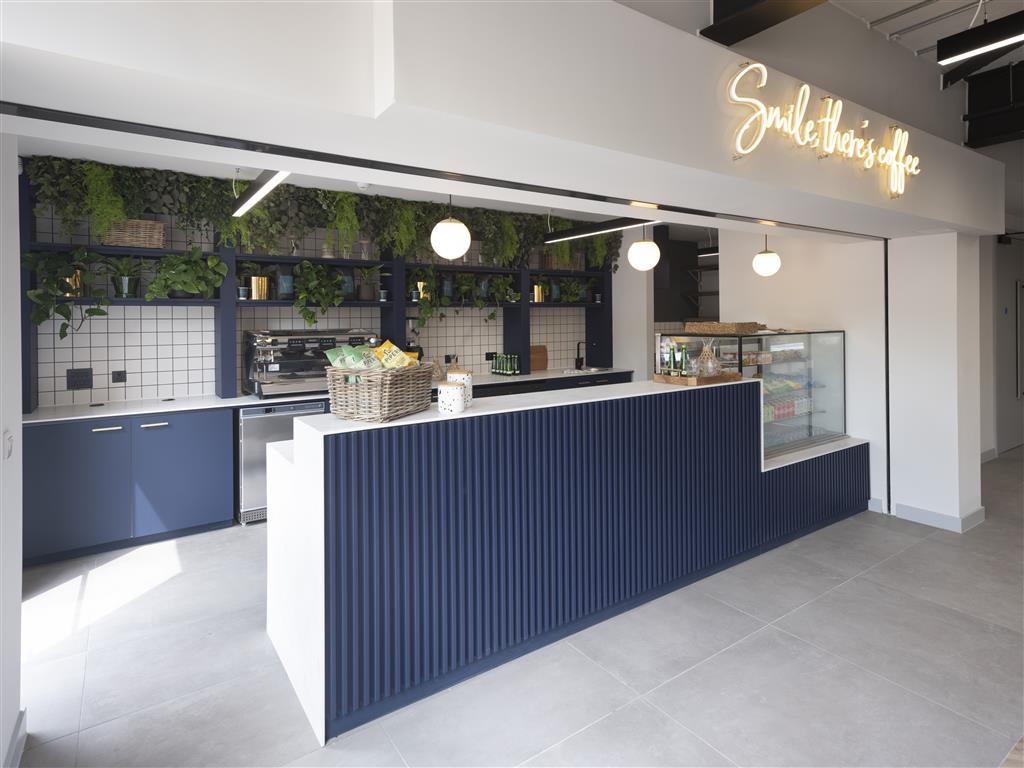 A modern coffee shop interior featuring a sleek counter with a ribbed blue facade. There’s a glass display case with pastries, hanging pendant lights, and a neon sign reading Smile, theres coffee. The verdant back wall adds a fresh touch of greenery.