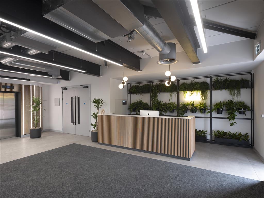 The modern office reception area features a wooden front desk, white computer, and hanging pendant lights. Verdant displays of greenery adorn the walls and shelves. Exposed ceiling ducts add character, while a lift and potted plant enhance the entrances welcoming vibe.