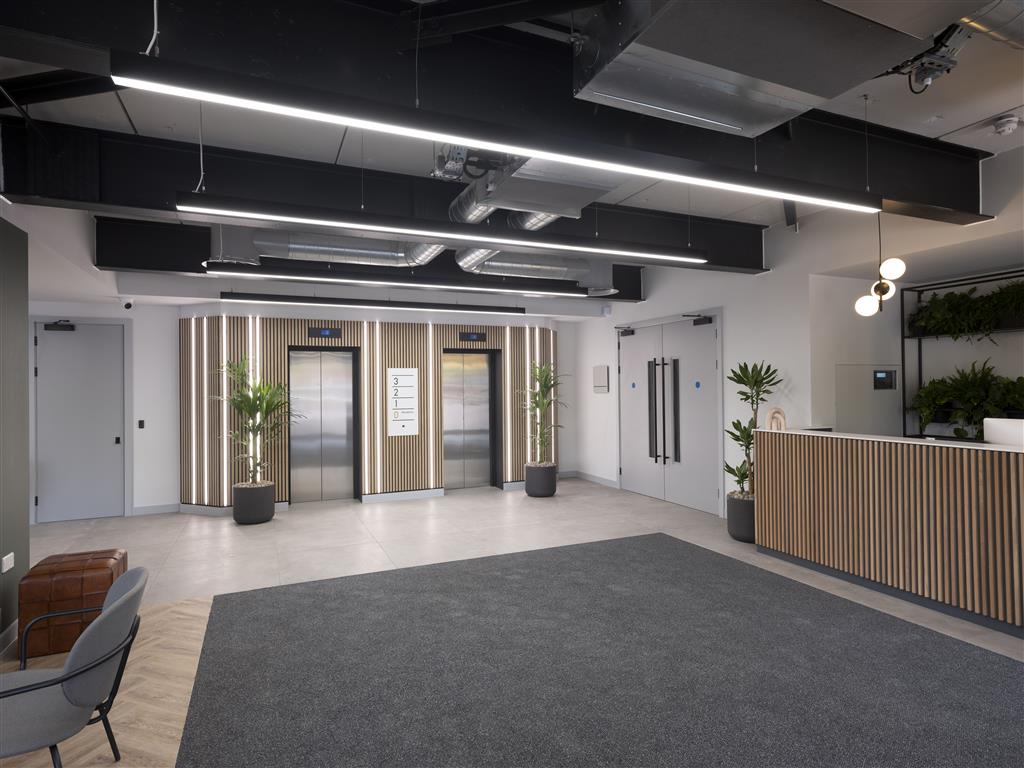The modern office lobby boasts two elevators and wood panel accents, with a verdant touch in the form of potted plants. A reception desk sits to the right, while a leather chair adds decor. Sleek overhead lighting complements the exposed ductwork, and light neutral tones grace the walls and floor.