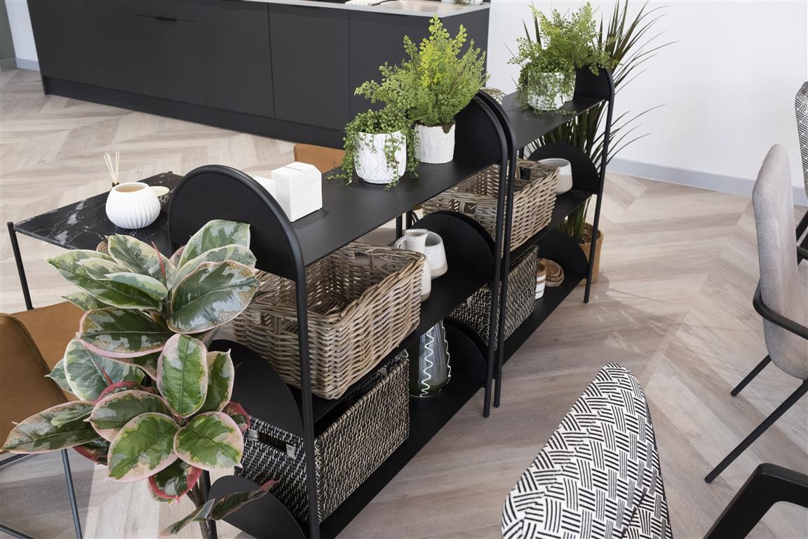 A modern, chic room with verdant touches features herringbone-patterned wooden flooring and a black shelf unit. The shelves hold wicker baskets, green plants, and decorative vases. Nearby, a marble-top table and stylish chairs enhance the contemporary decor.