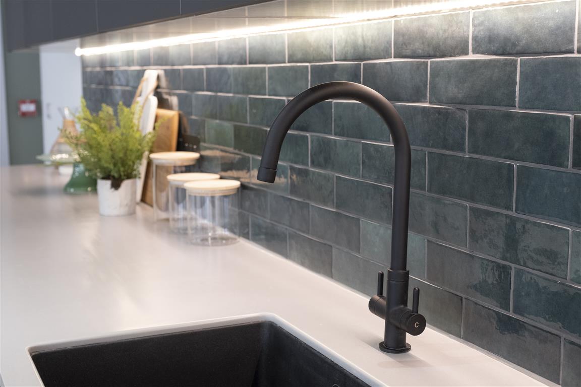 A modern kitchen sink with a matte black faucet is set against a backsplash of verdant, glossy dark green tiles. The countertop features potted plants, a cutting board, and sealed jars. Under-cabinet LED lighting illuminates the area.