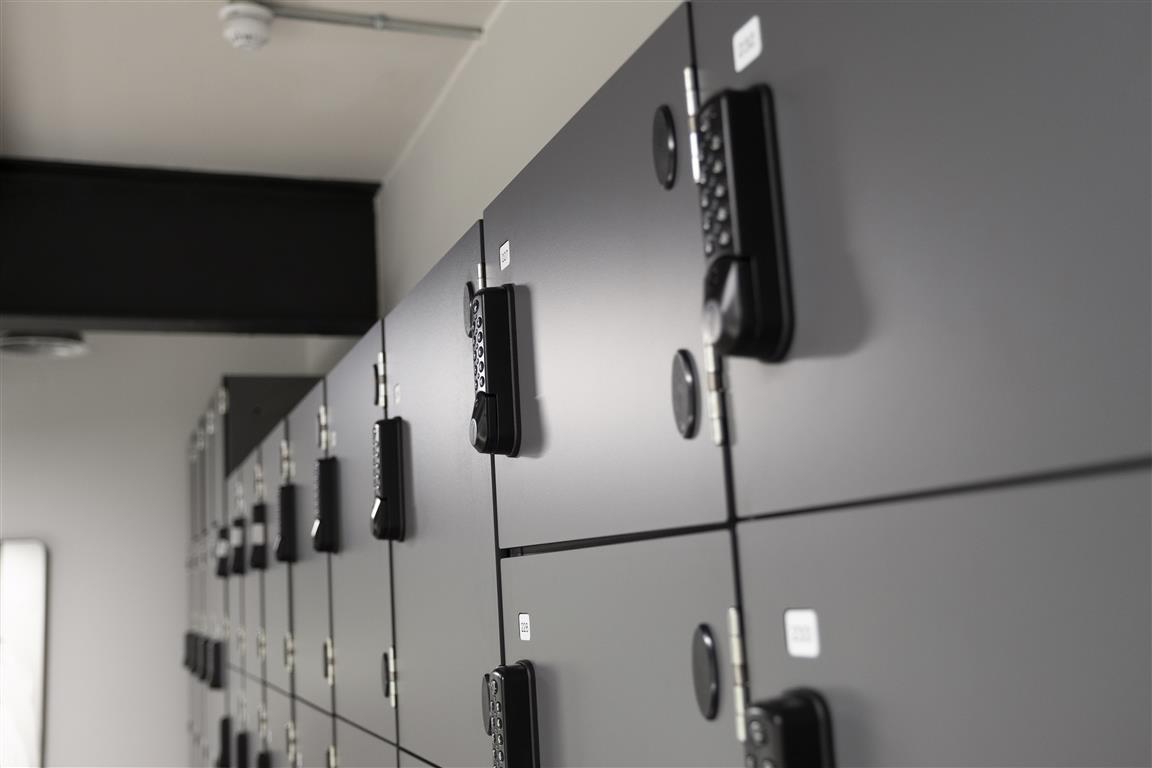 A row of black lockers with electronic combination locks is viewed at an angle. The sleek, modern design, complemented by labeled sections, exudes a minimalist verdant aesthetic amidst the rooms light ceiling and visible ventilation pipes.