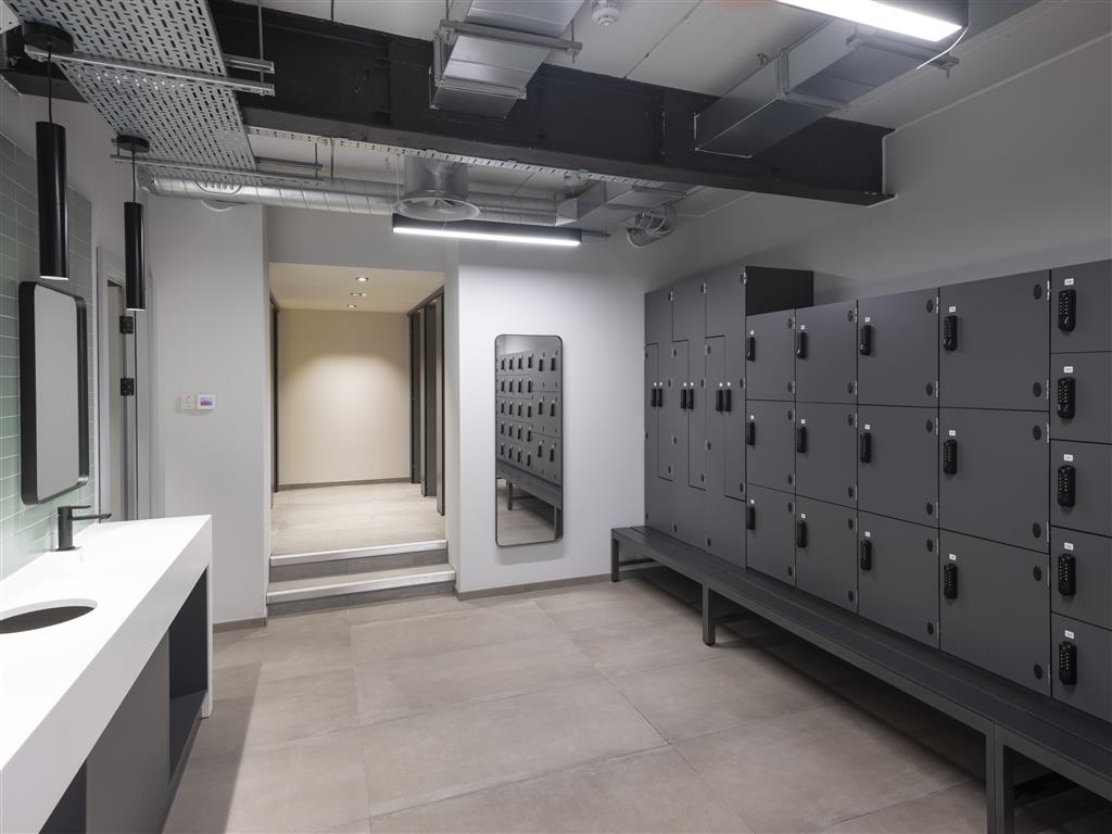 A modern locker room with gray lockers and a bench on the right. Verdant touches bring life to the space. A large mirror is mounted on the wall, while the left side boasts a white countertop with sinks and mirrors. Overhead lighting and exposed ducts lend an industrial feel.