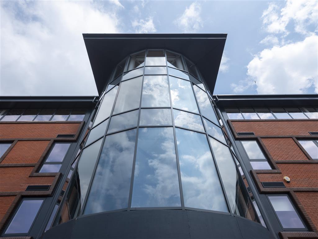 Architectural perspective of a modern building with expansive, reflective glass windows set against a cloudy sky. The structure boasts verdant surroundings, red brick walls, and a prominent central glass tower that mirrors the clouds above.