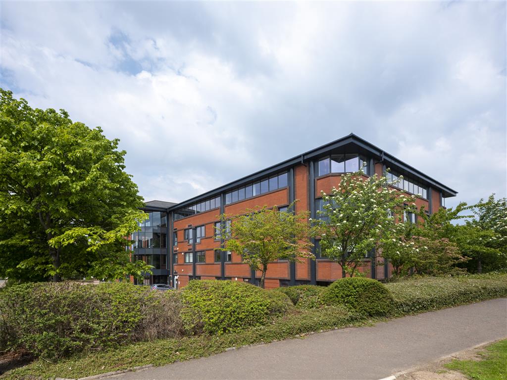 A modern red-brick building with large windows stands amidst verdant trees and shrubs under a partly cloudy sky. A paved pathway runs alongside the building.