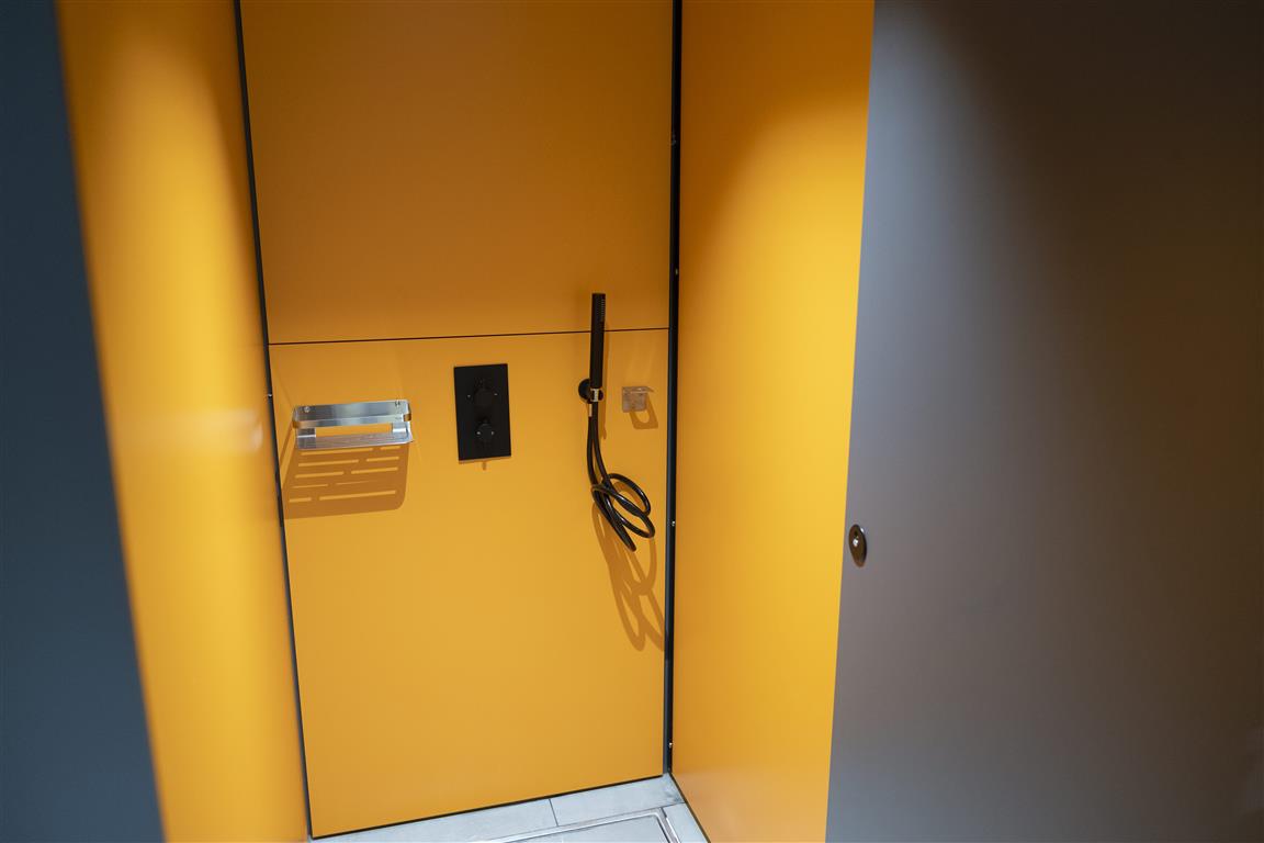 The yellow locker, contrasted by verdant surroundings, features a black charging cable and metal shelf inside. Its door is ajar, revealing the interior setup, while the gray area with chrome accents frames its vibrant presence.