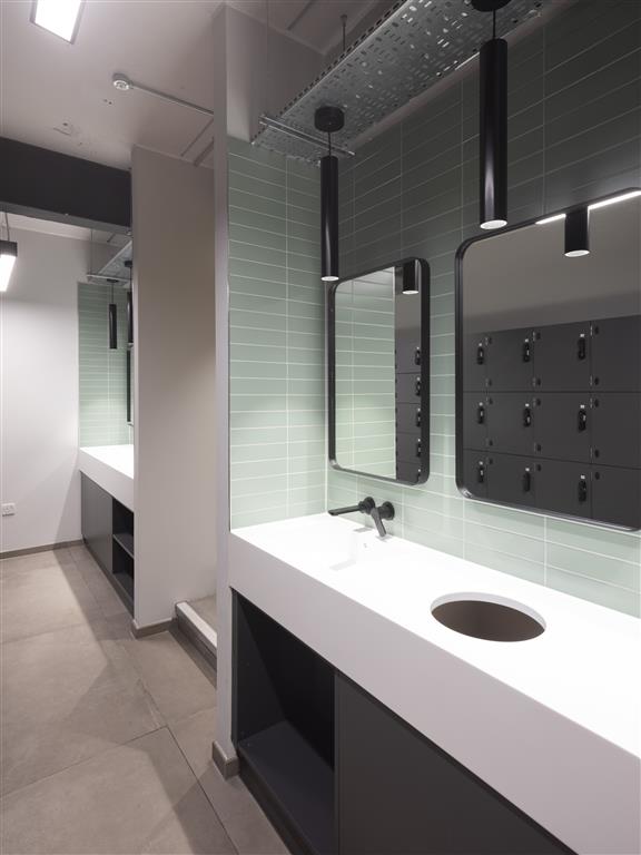 Modern restroom with verdant tiled walls and two sinks. Rectangular mirrors hang above each sink. Black faucets and pendant lights complement the sleek design. Locker units are visible on the right. Neutral tones create a minimalist look.