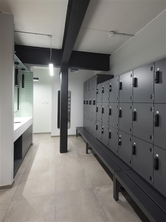 The modern locker room features rows of grey lockers and a bench under bright overhead lights. A verdant touch adds freshness to the space, with plants near the sink area and mirrors. The light-colored tile floor completes this inviting setting.