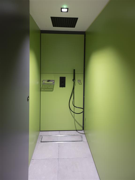 A modern shower area with verdant walls, featuring a black handheld showerhead and a square overhead shower. The ceiling has a dark panel with a small green light, and the floor is light gray with a drainage grate.