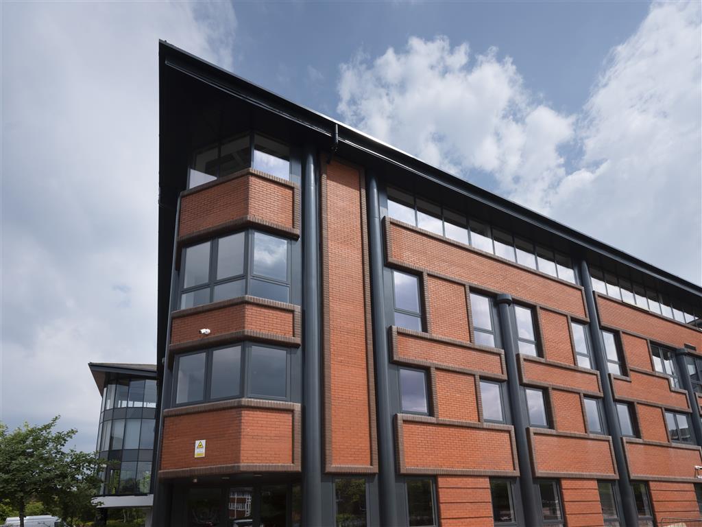 Verdant trees frame the lower left of this modern building, which boasts a red brick facade and large, dark-framed windows. The structure features angular design elements, set against a partly cloudy sky.