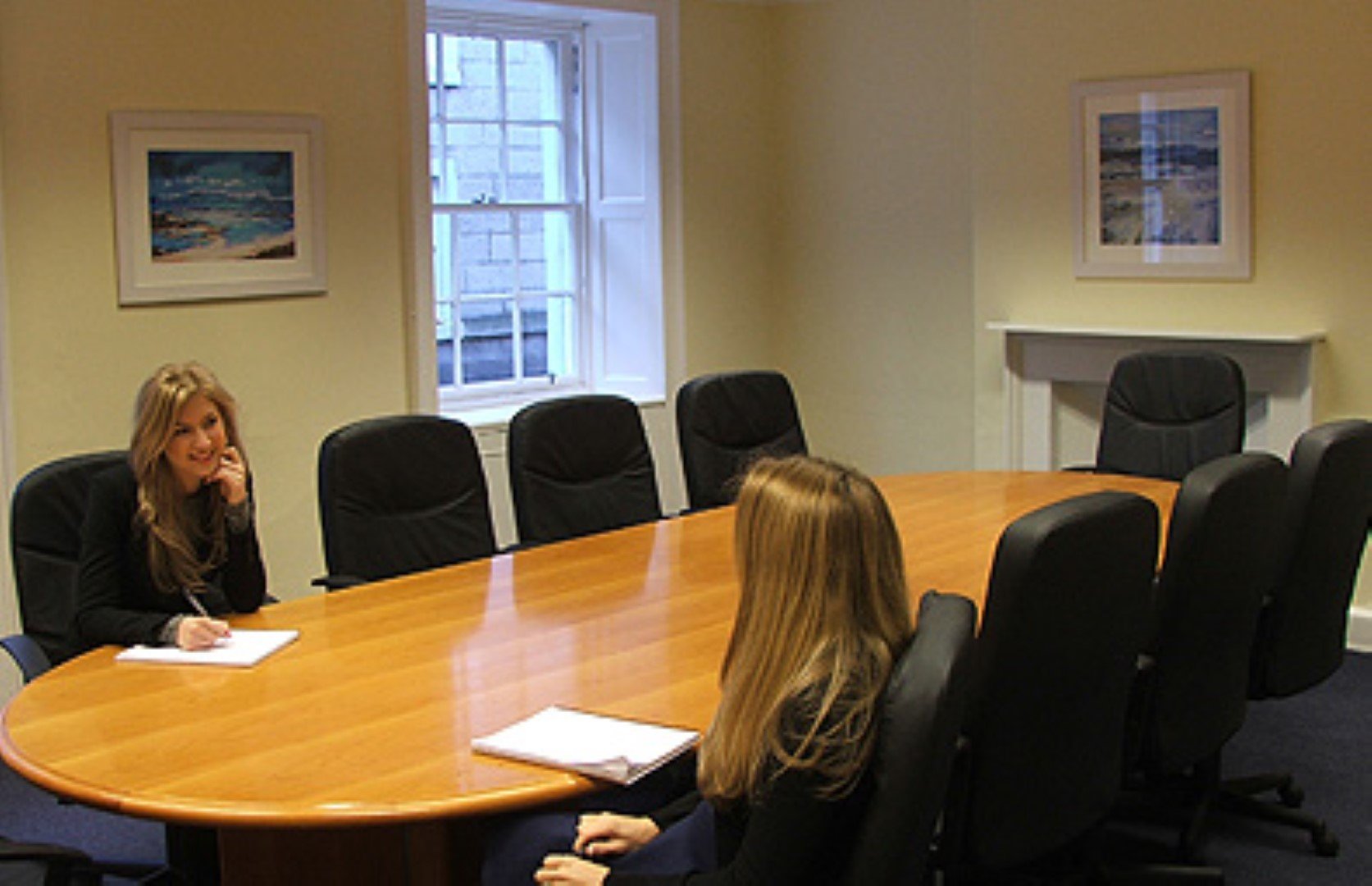 Two individuals sit at opposite ends of a long wooden conference table in the Hill Street meeting room, surrounded by black chairs and white walls adorned with two paintings. One person is writing; both have long hair. A window lets in natural light.