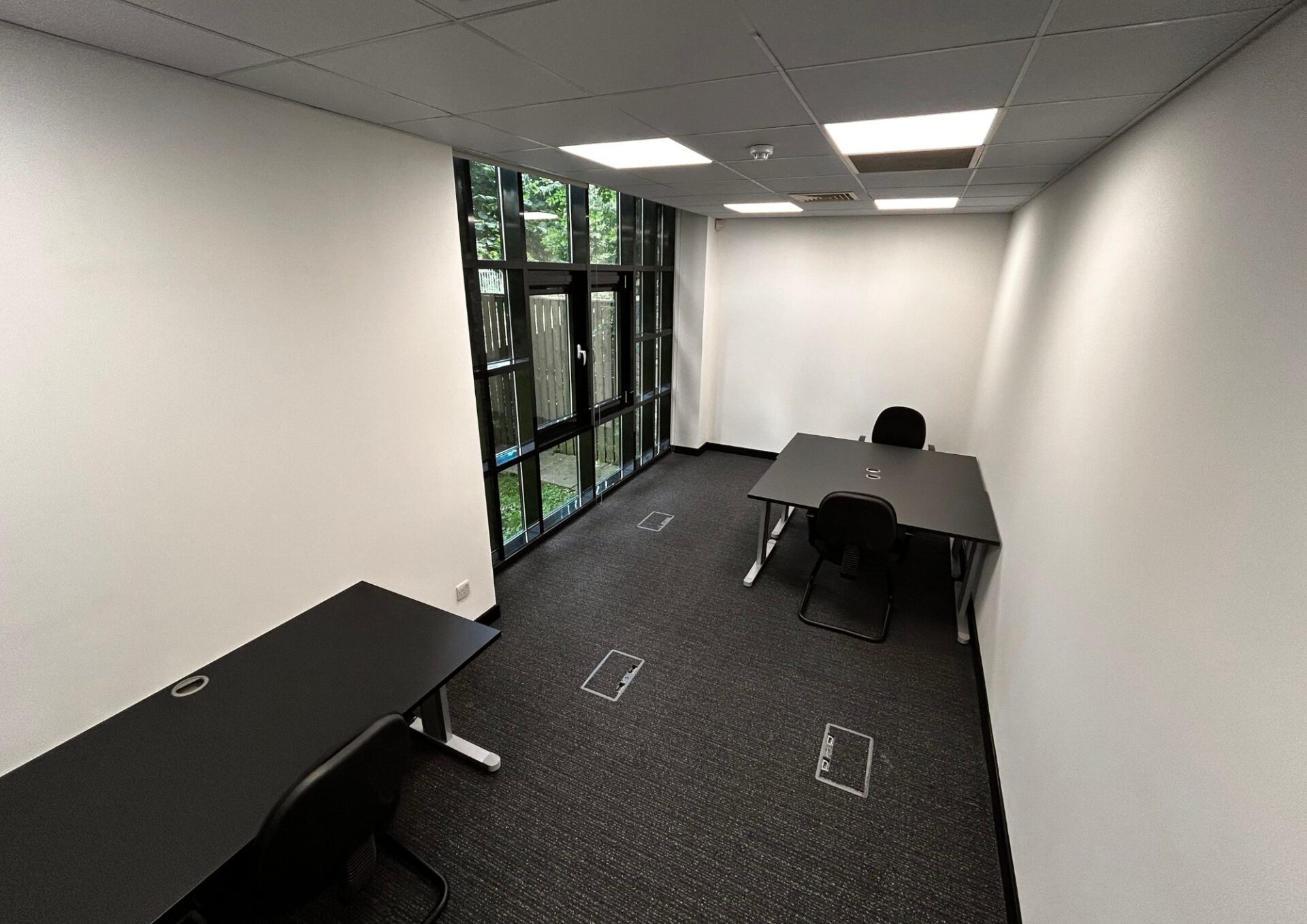Office 1 Papermill Wynd features a narrow room with three black desks and matching chairs. White walls and a glass door complement the large window on the left, allowing natural light to stack effortlessly upon the dark gray carpeted floor.