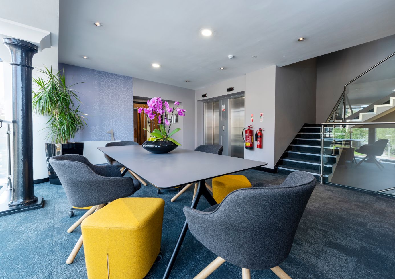 The modern office lobby in Edinburgh Leith features a long table surrounded by gray chairs and two yellow stools. A vase with pink orchids graces the tabletop. In the background, two elevators sit beside a staircase with glass railing, bright lighting, and blue carpet complete the scene.