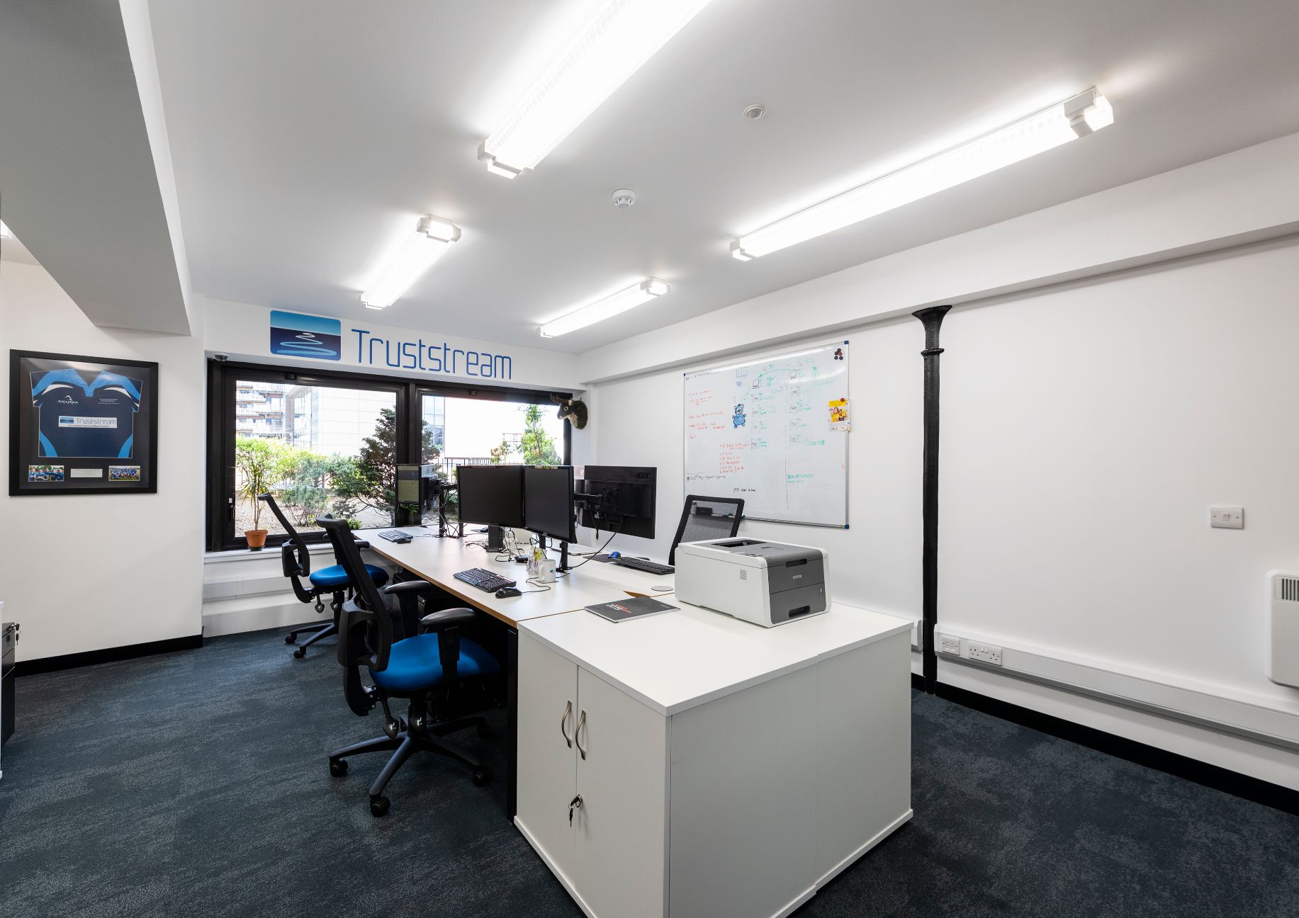 Modern office space in Edinburgh with white walls featuring a sign reading Truststream. Located in Leith, it has desks with computers and chairs, a copier, and a whiteboard on the wall. Large windows in the back offer natural light and a view of greenery outside.