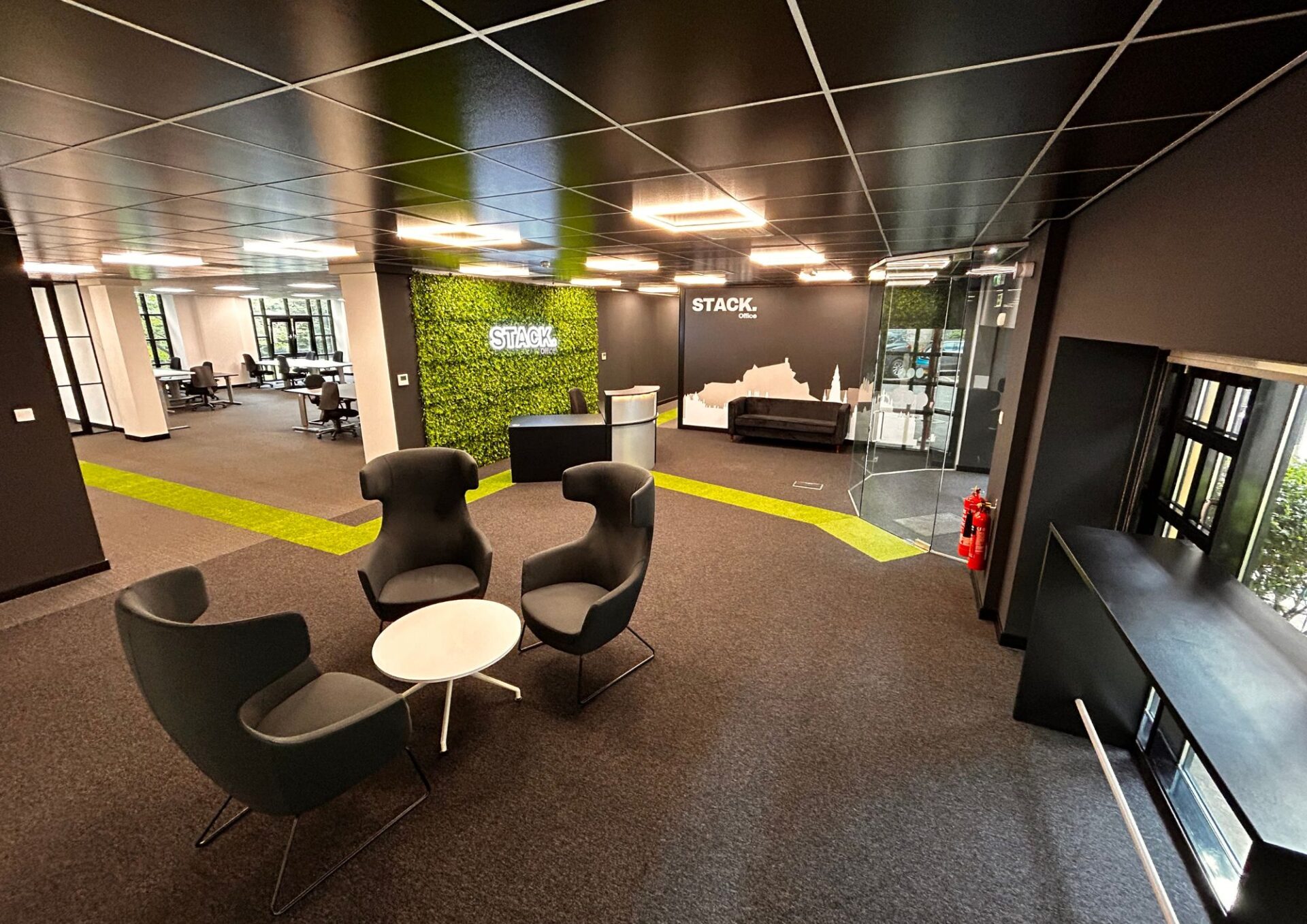 Modern office lobby at Office 1 Papermill Wynd features black walls and ceiling, a green accent wall with a STACK logo, and four black chairs around a white table. The open workspace in the background has desks and computers. Bright lighting and a green strip on the carpet enhance the decor.