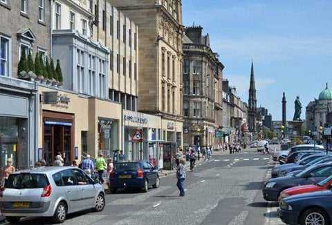 A busy street in Edinburgh with parked cars on both sides. People stroll along the sidewalks near charming shops and tall historic buildings. A distant spire and statues lend a classic touch under the clear blue sky.
