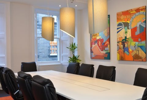 A modern conference room in Edinburgh features a long white table and black chairs. Abstract colorful paintings adorn the walls, while large pendant lights hang from the ceiling. A leafy plant by the window invites natural light into this stylish space.