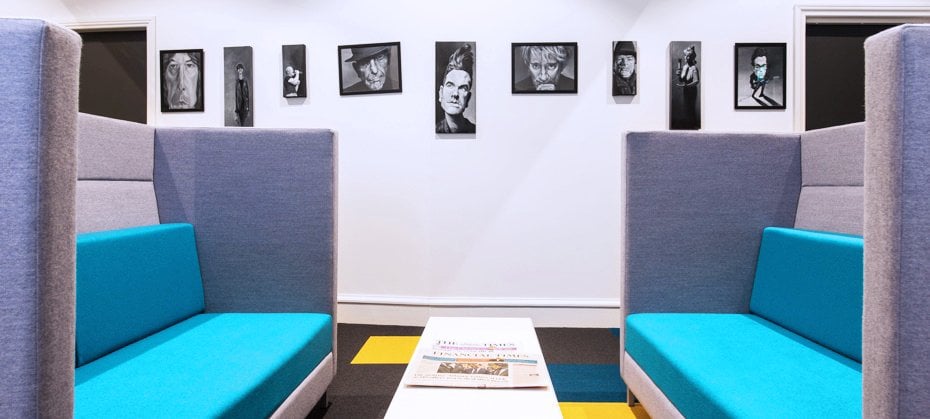 A modern seating area in Edinburgh features two gray booths with turquoise cushions, separated by a white table. Black and white portraits adorn the wall, while the floor showcases a geometric pattern in yellow, black, and gray.