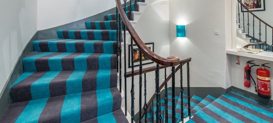 A spiraling staircase with blue and gray striped carpet, wooden banister, and black railing, reminiscent of an elegant Edinburgh townhouse. It winds around a white wall adorned with framed artwork and turquoise wall lights. Two fire extinguishers are mounted on the wall near the stairs.