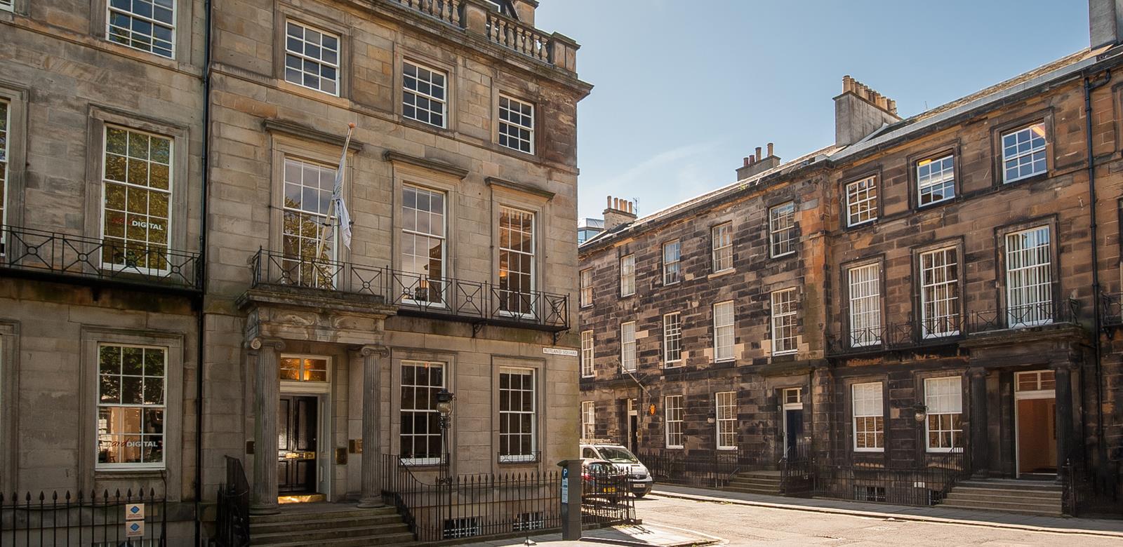 A quiet street with historic stone buildings featuring traditional sash windows and wrought iron railings. The architecture showcases carved stone details. A parked car is visible, with clear skies above.
