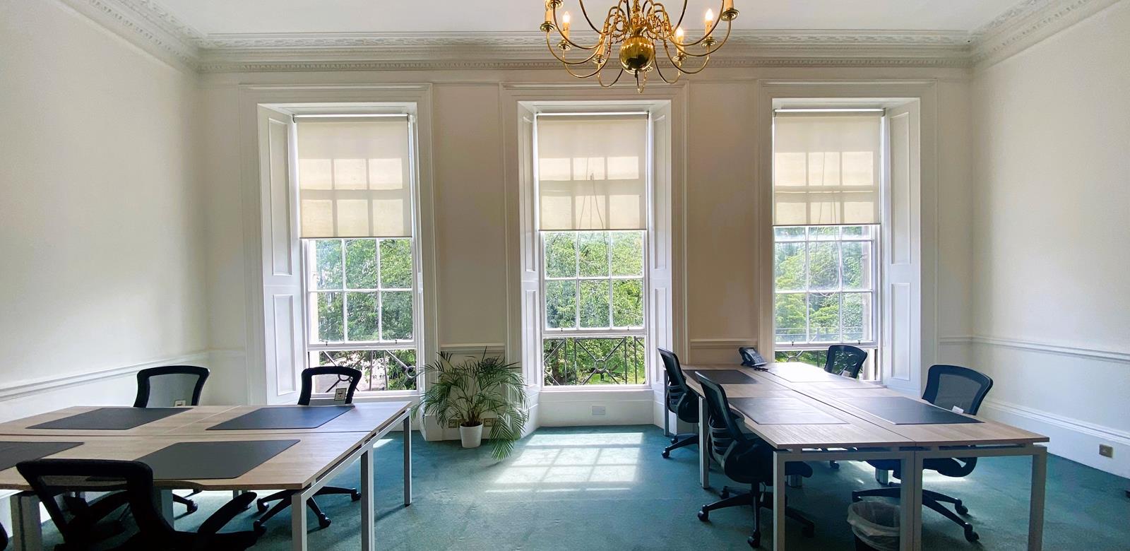 Bright office space with large windows, two desks, chairs, and a potted plant. Sunlight streams in, illuminating the light green carpet and white walls. A decorative chandelier hangs from the ceiling.