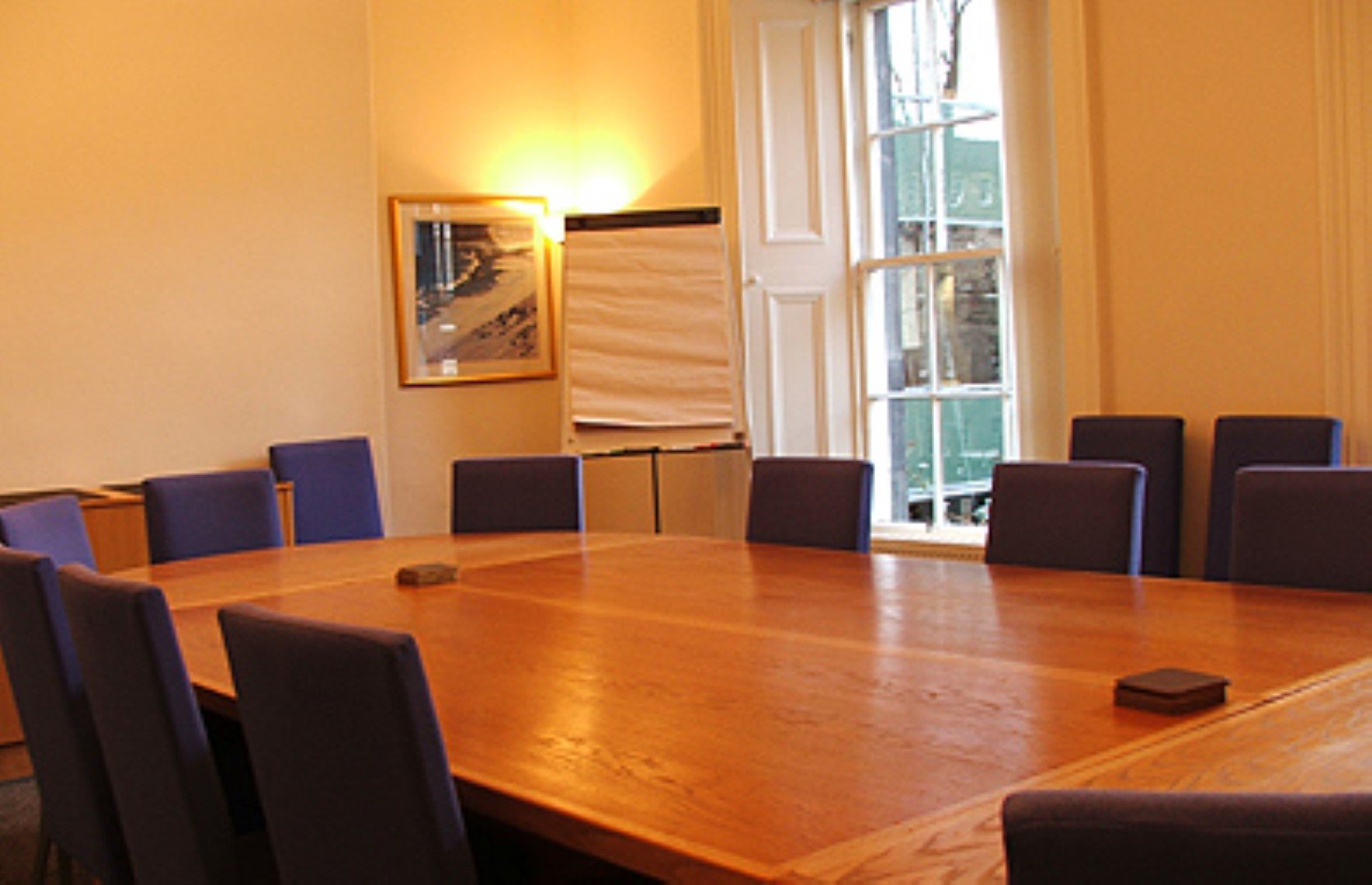 The conference room at Thistle Court Business Centre features a large wooden table surrounded by blue chairs. An easel with blank flip chart paper stands near a window, while a framed picture adorns the wall, all illuminated by natural light streaming through the tall window.