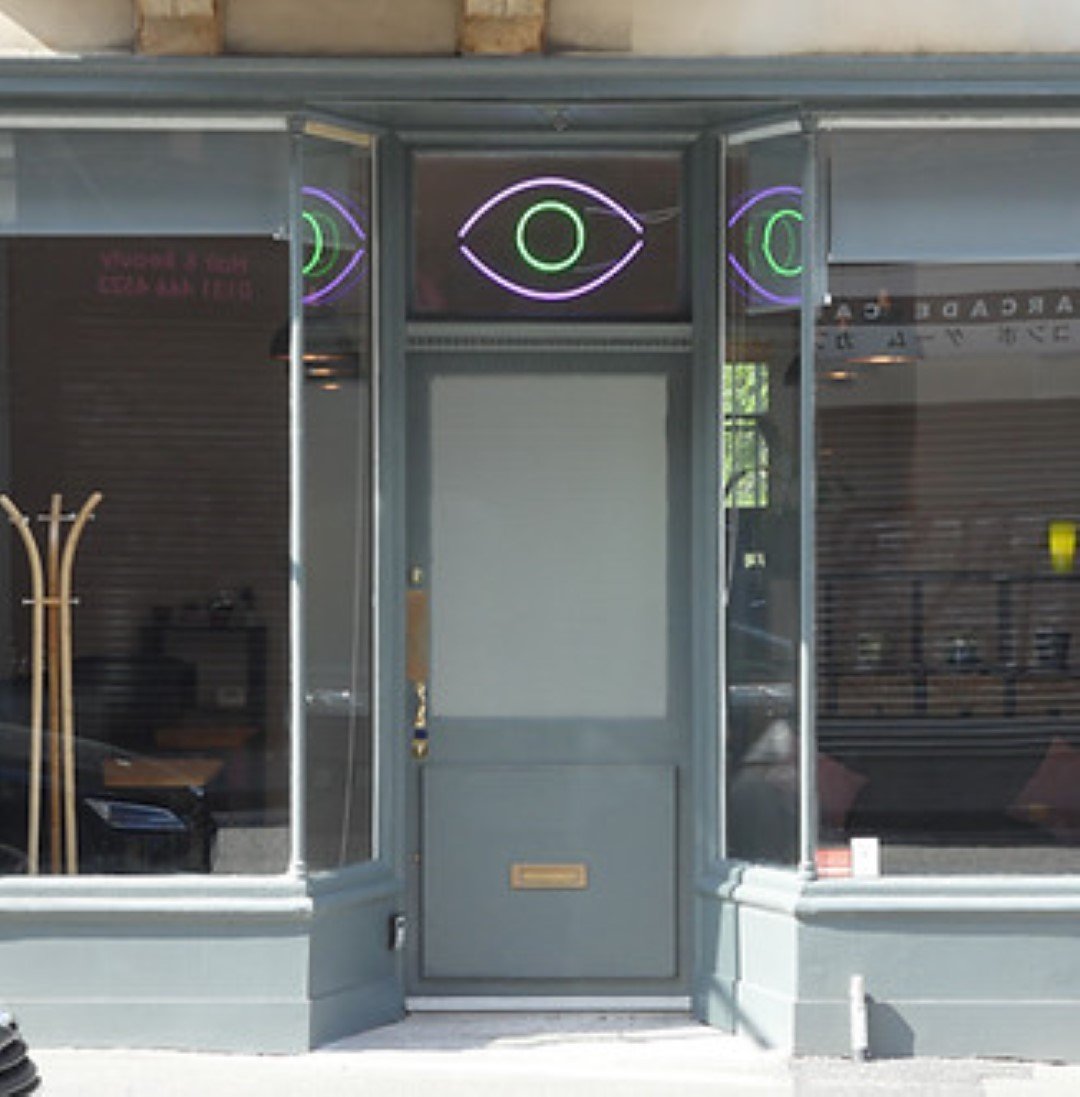 The shopfront boasts a gray door and expansive glass windows. Above, a neon sign of an eye subtly hints at the mysteries within The Nexus Room. Inside, a coat rack and chairs are visible through the windows, inviting curious onlookers to explore further.