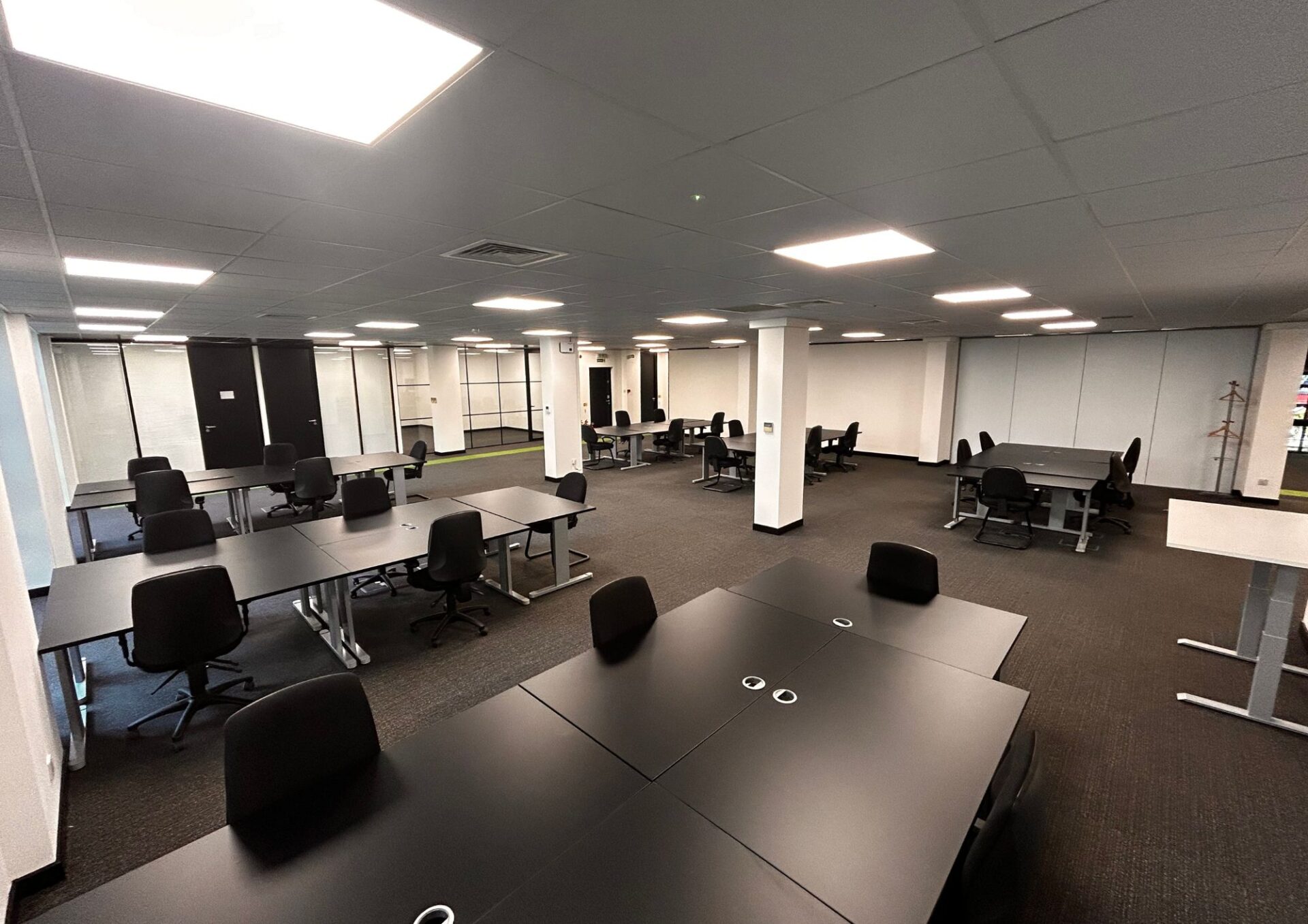 Office 1 Papermill Wynd boasts a spacious interior with STACKed arrangements of sleek black desks and office chairs. The room is brightened by overhead lights, features a dark carpeted floor, and has crisp white walls, with columns and dividers elegantly scattered throughout.