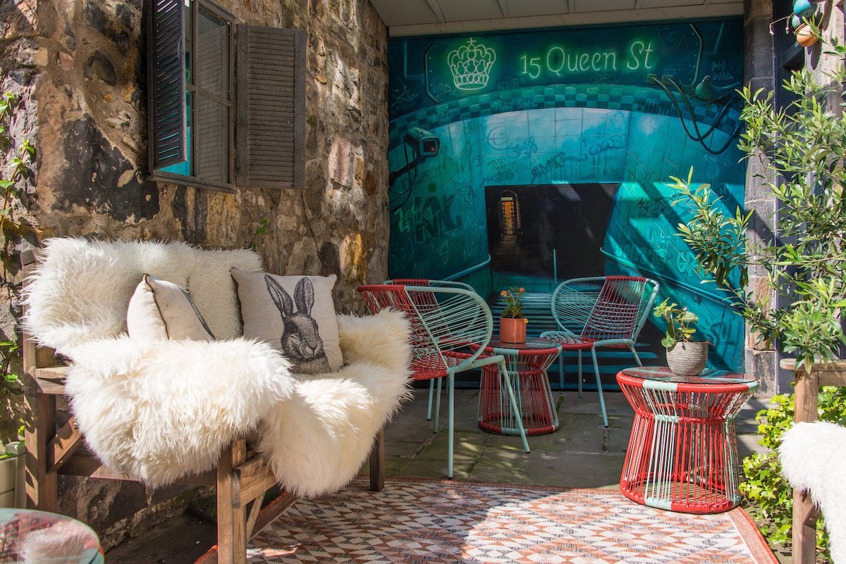 A cozy outdoor seating area with a fluffy white bench adorned with pillows, including one with a rabbit design. Red and blue wire chairs and tables sit beneath a mural reading 15/16 Queen Street along with a crown design. Decor includes a small potted plant.