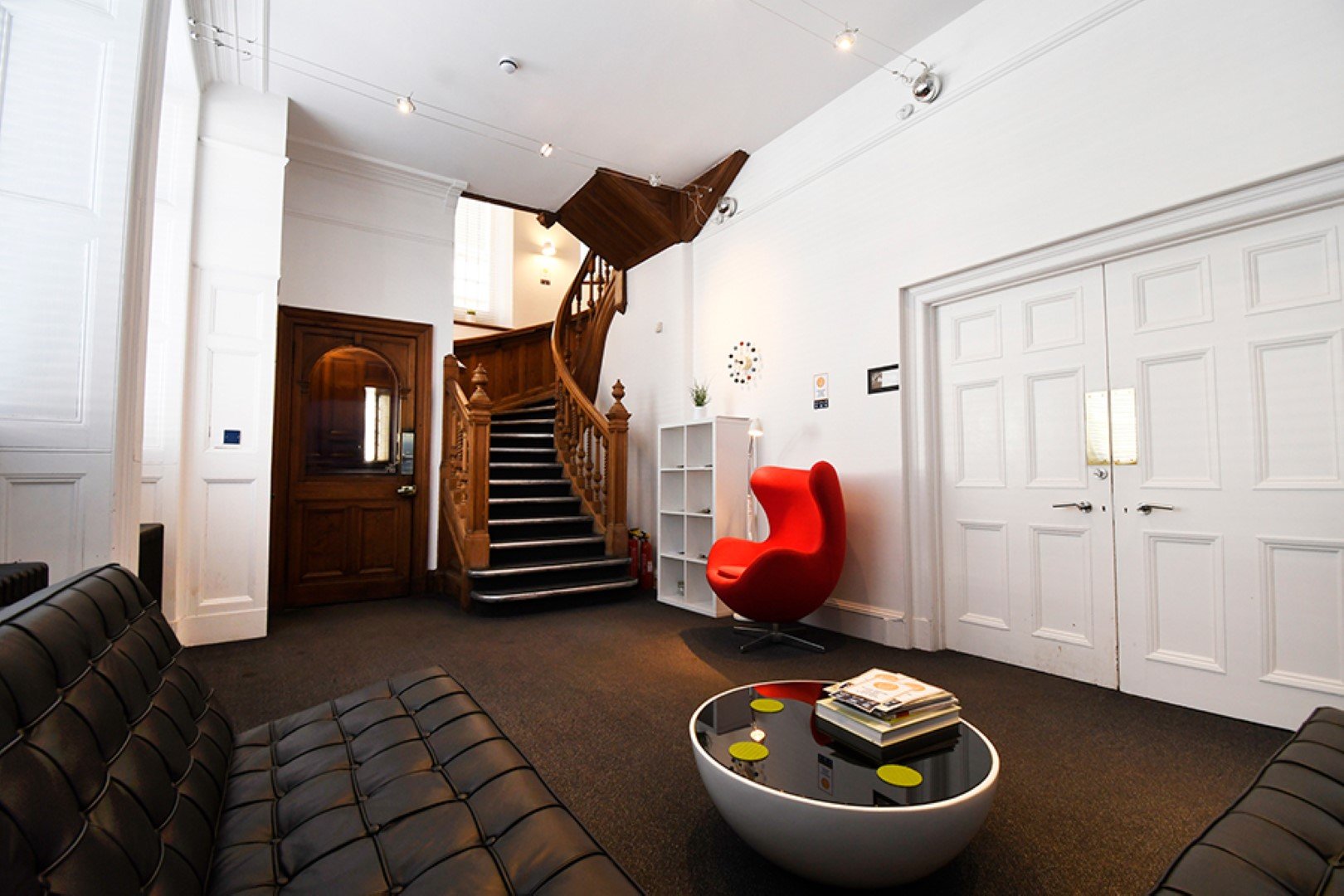 Modern lounge with dark sectional sofas, a red egg chair, and a round coffee table featuring colorful coasters from 93 Unlimited. A wooden staircase ascends near a closed wooden door. White walls and double-door entrance add to the spacious, minimalist feel.