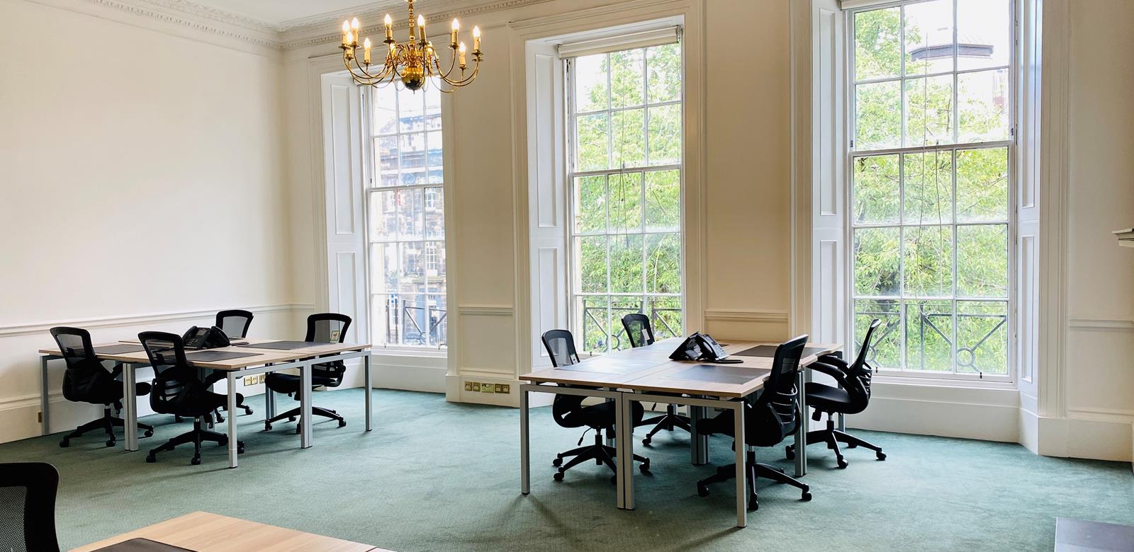 A bright, spacious office room with three wooden desks, each paired with black office chairs. Large windows overlook green trees, and a chandelier hangs from the ceiling. The carpet is light green.