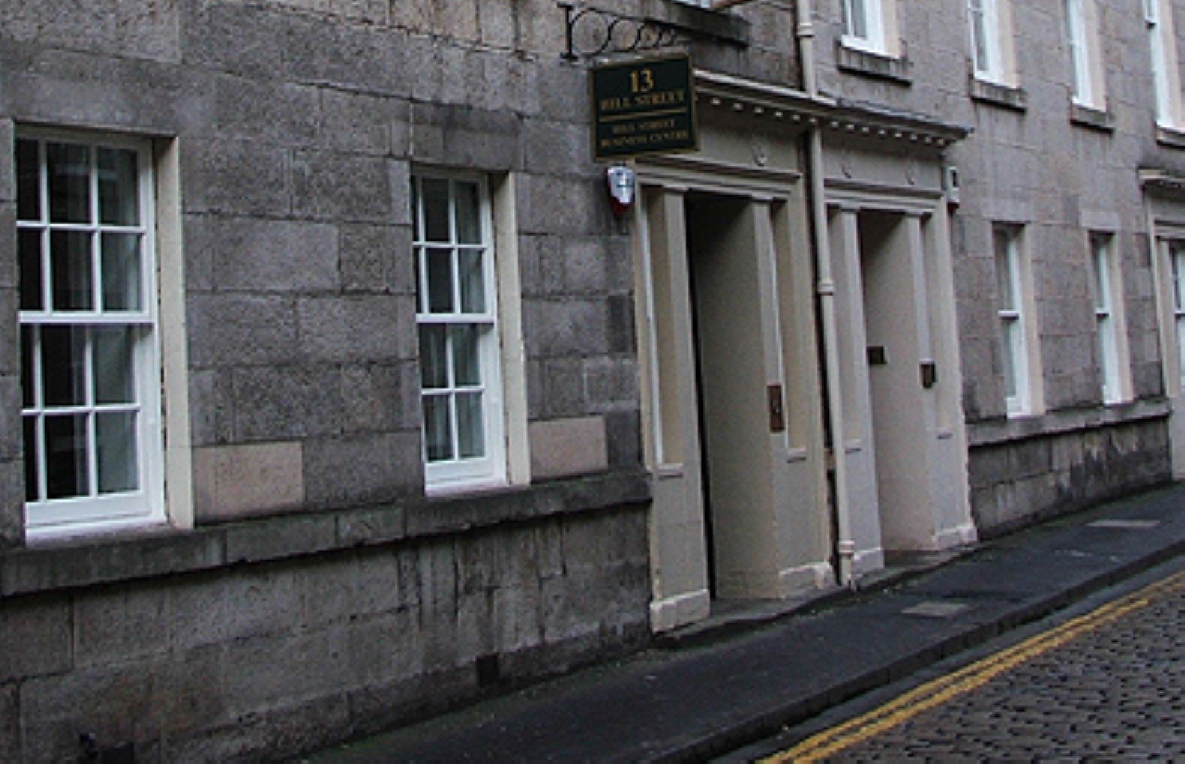 A narrow, empty street with stone buildings stands silent. One building proudly showcases a sign above the doorway reading 13 Hill Street. The gray walls contrast with the white window frames, while the cobblestone-lined sidewalk whispers stories of history and charm.